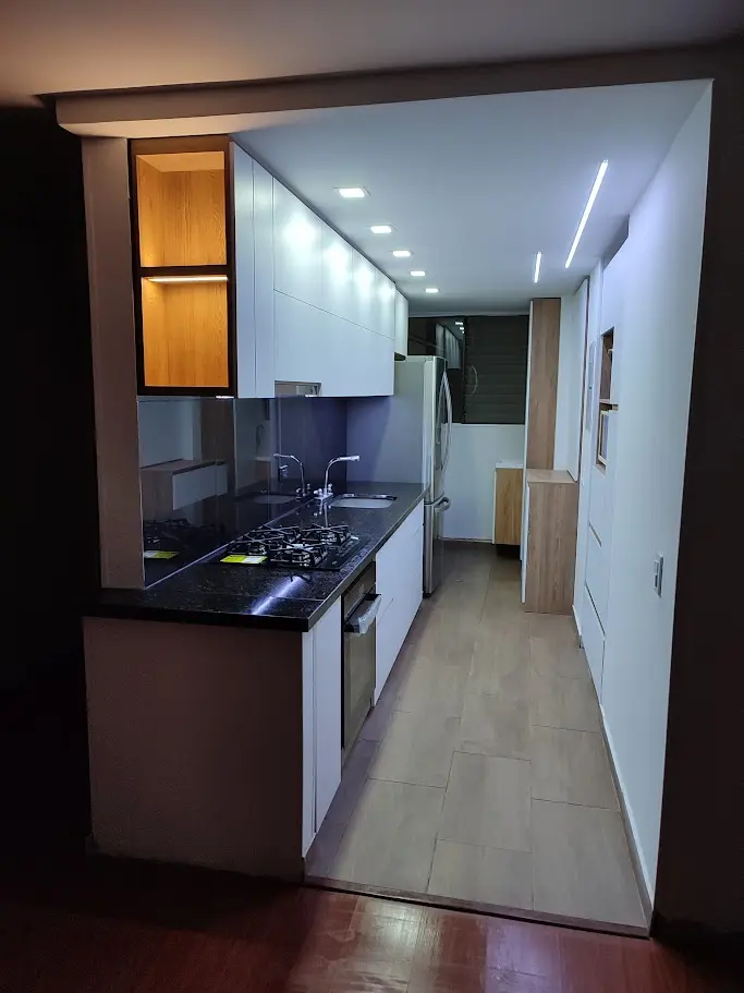 Diseño de cocina integral lineal con muebles blancos, mesón en granito negro e iluminación LED cálida